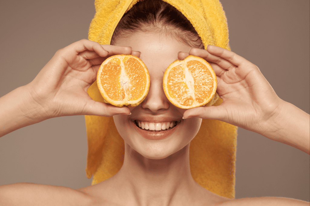 image featured in How To Make Your Skin Care Routine Sustainable in 7 Easy Ways showing a smiling woman holding oranges to her eyes